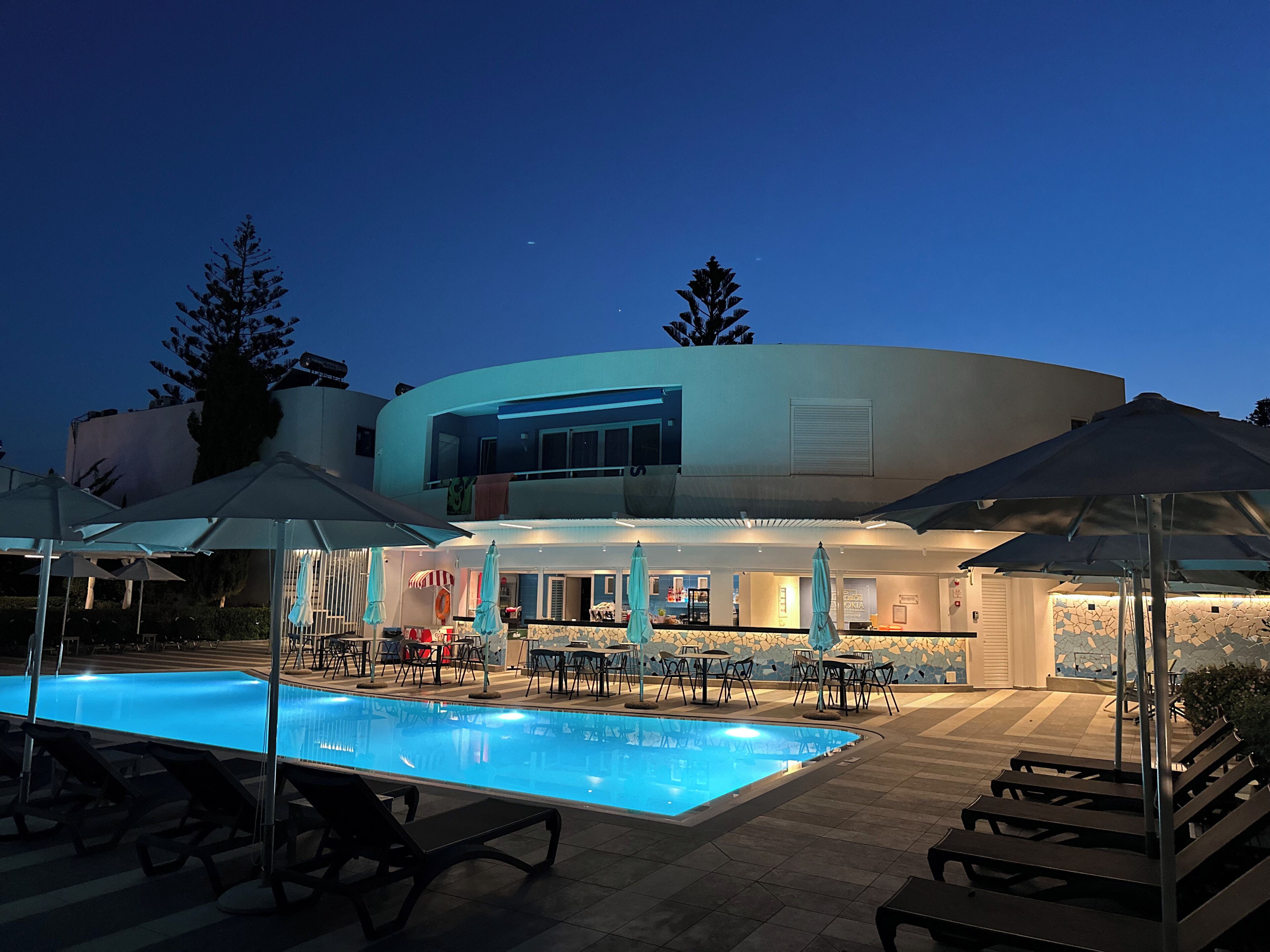 A pool area with lounge chairs and umbrellas, a bar, and a building with a curved facade.