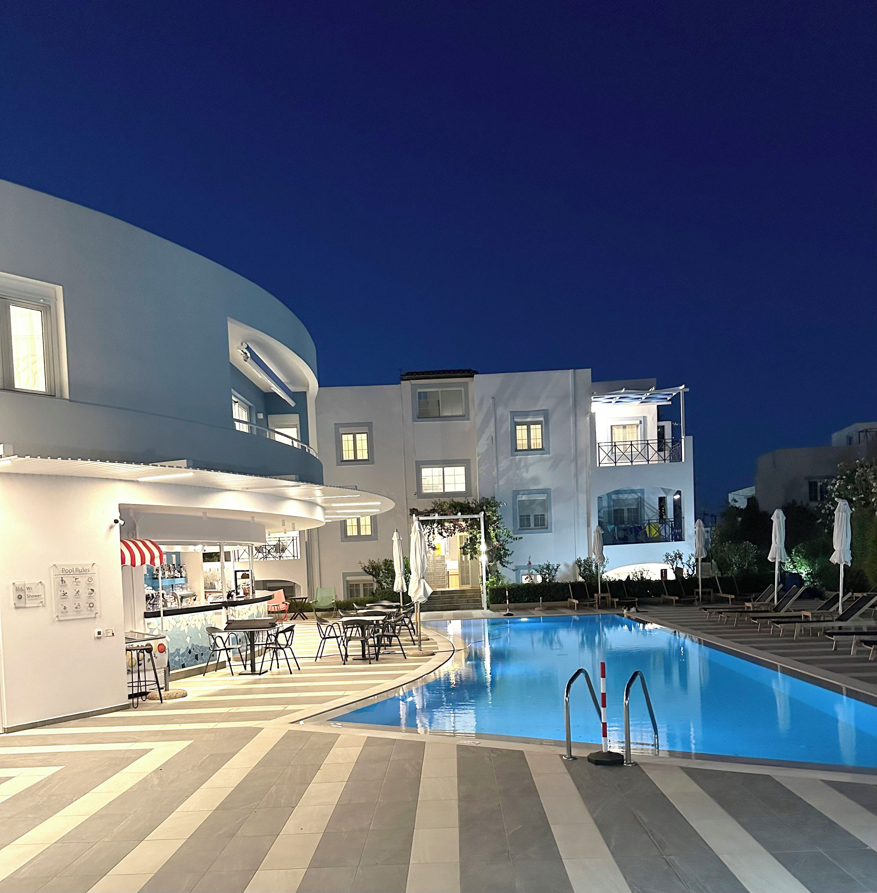 An indoor swimming pool with lounge chairs and tables, surrounded by modern buildings with balconies and large windows.
