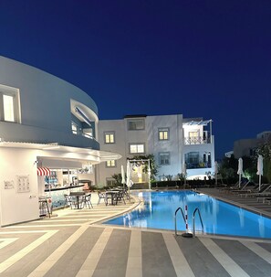 An indoor swimming pool with lounge chairs and tables, surrounded by modern buildings with balconies and large windows. - Evdokia Suites (Hersonissos)