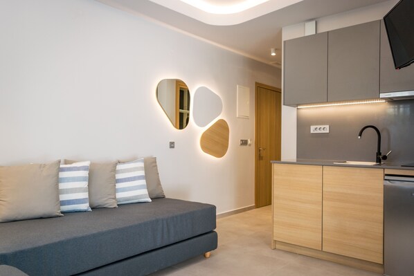 A modern living room with a grey sofa, wooden cabinets, and a built-in kitchen with a sink and stove. - Evdokia Suites (Hersonissos)