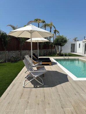 A poolside area with lounge chairs and umbrellas, a stone wall, and a pool. - Evdokia Suites (Hersonissos)