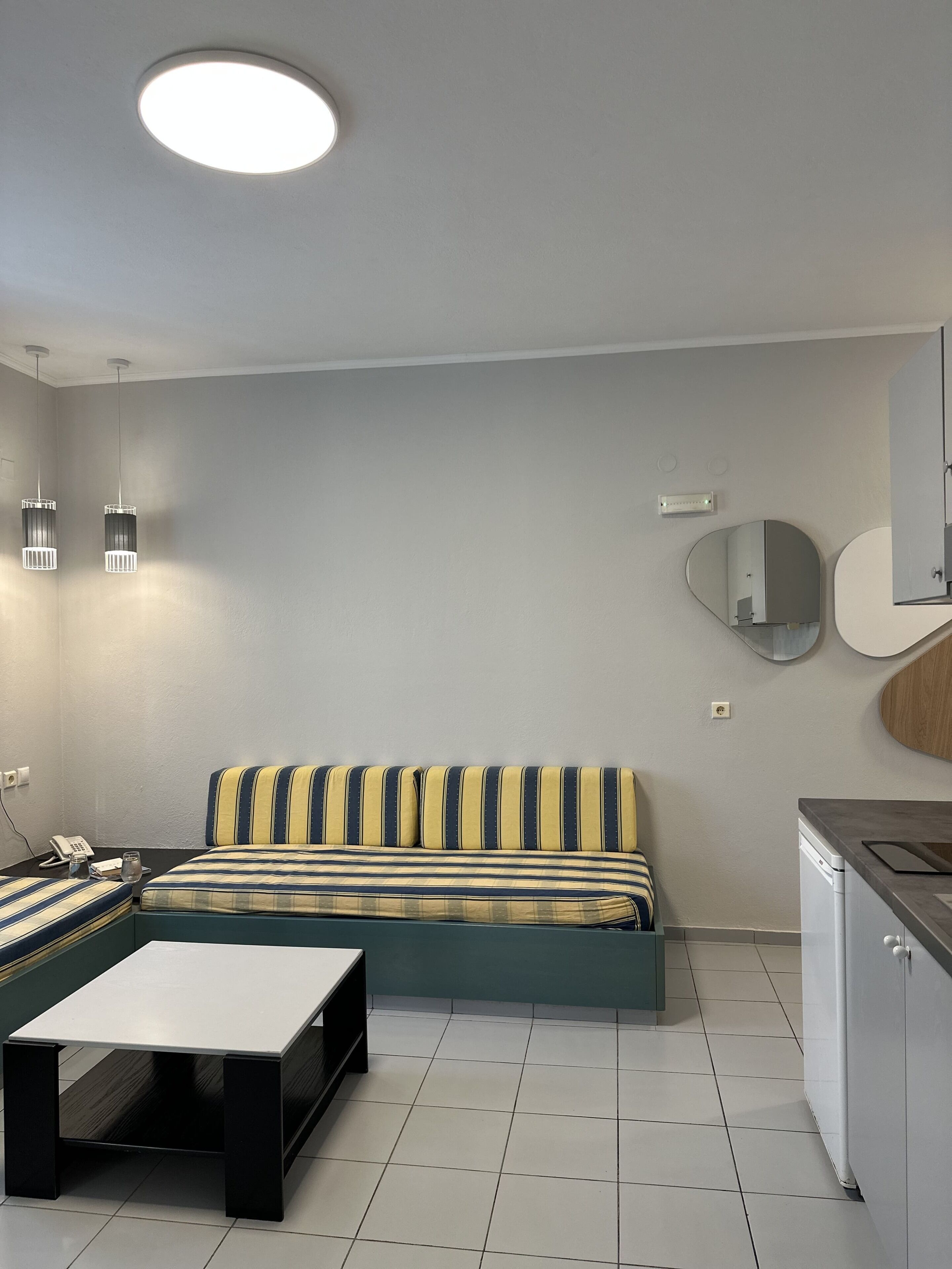 Een compacte woonruimte met een bank, een klein tafeltje, een keuken met een vaatwasser, en een plafondlamp.