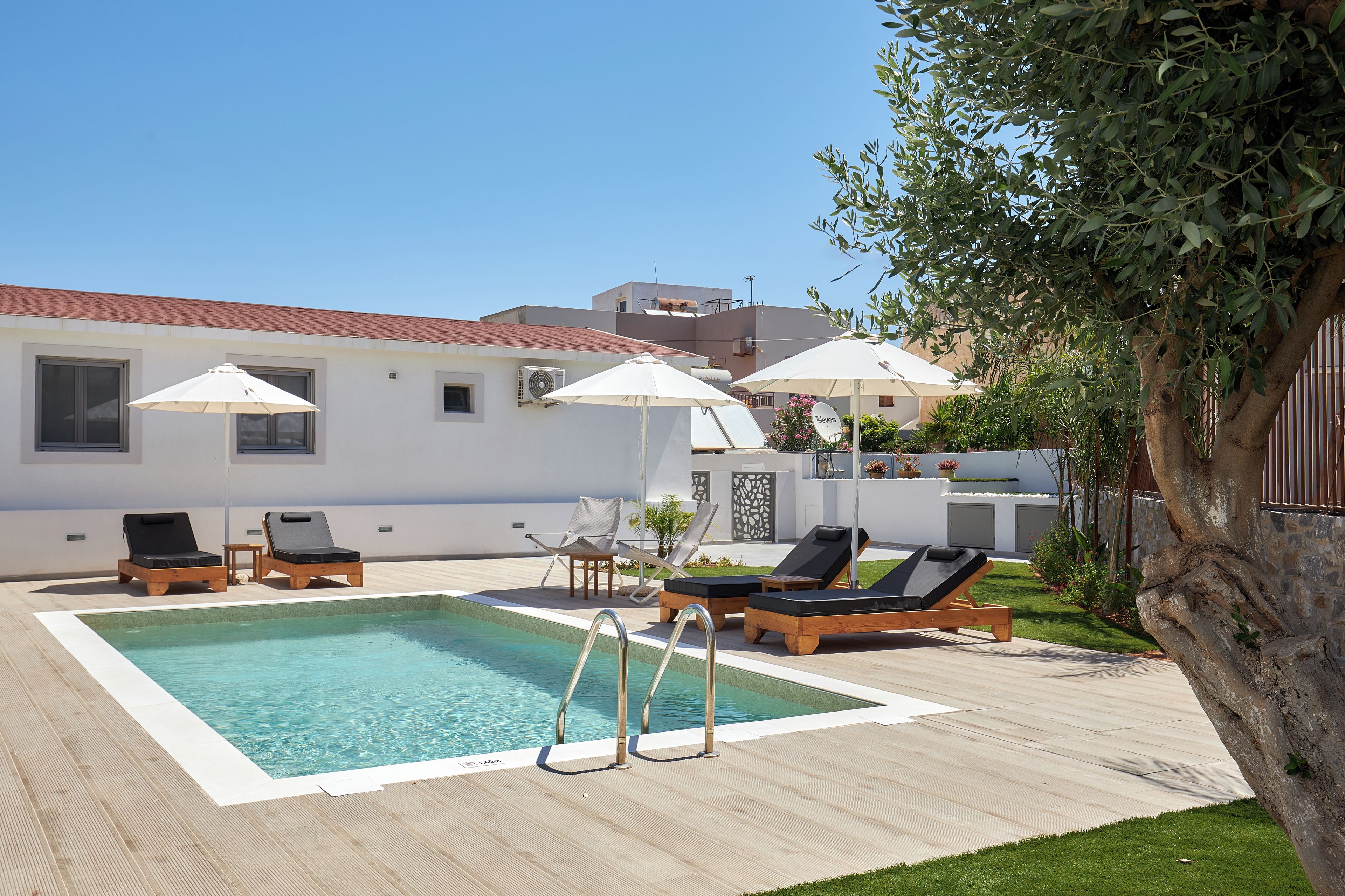 Une piscine entourée de chaises longues et de parasols, elle-même bordée d’un bâtiment blanc doté d’un toit en tuiles.