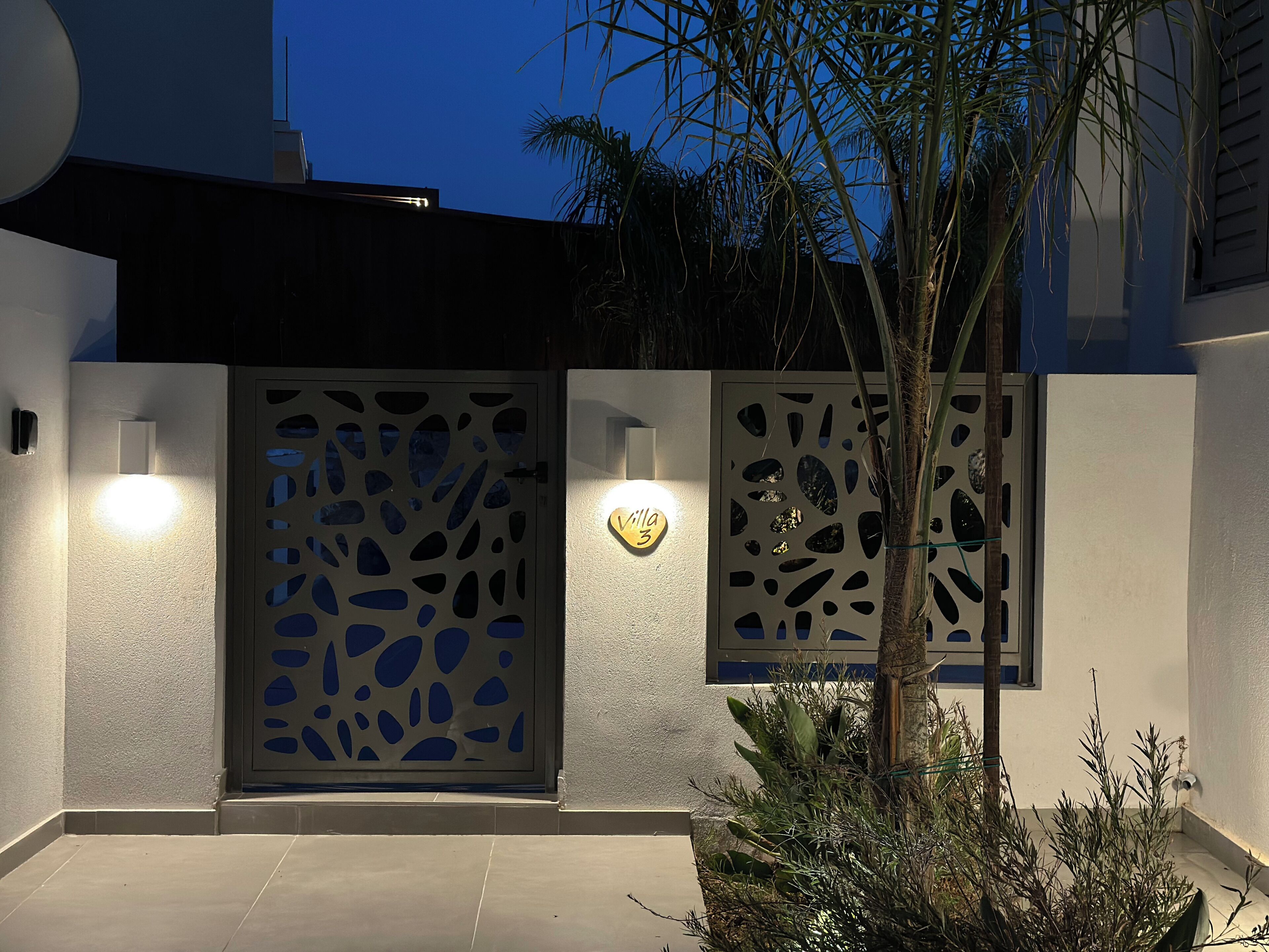 A modern building entrance with a decorative metal gate, a wall-mounted light, and a small tree.