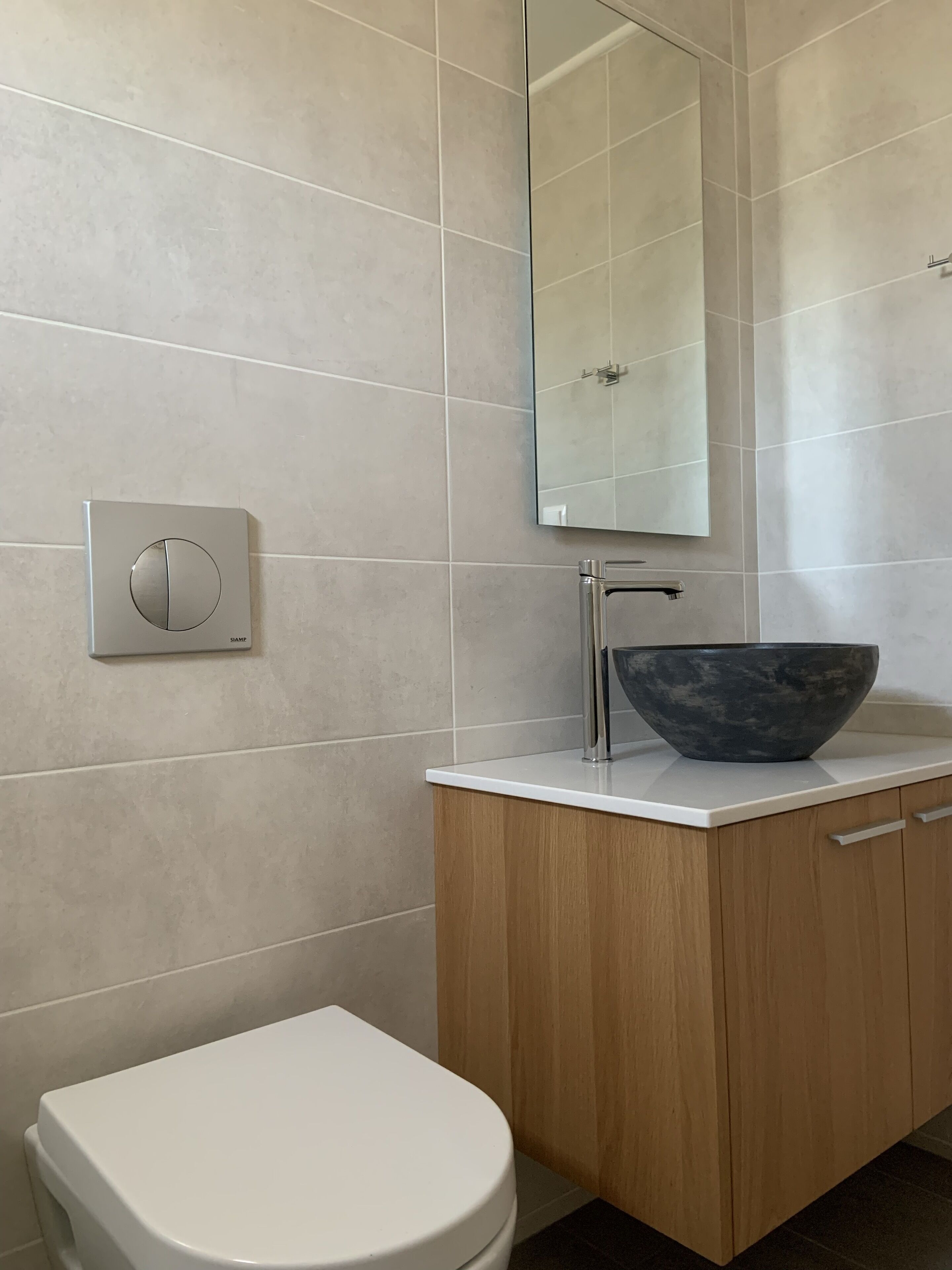 A modern bathroom with a toilet, a wooden cabinet, a white countertop, a large mirror, and a wall-mounted light switch.