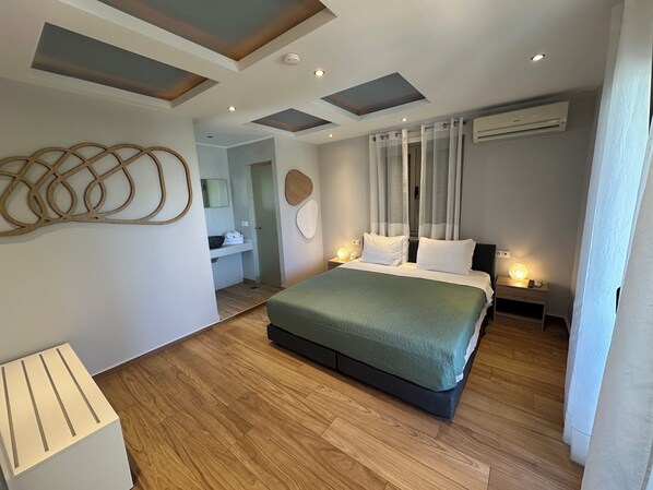 A modern bedroom with a large bed, a wooden floor, recessed ceiling lights, and a bathroom visible in the background. - Evdokia Suites (Hersonissos)