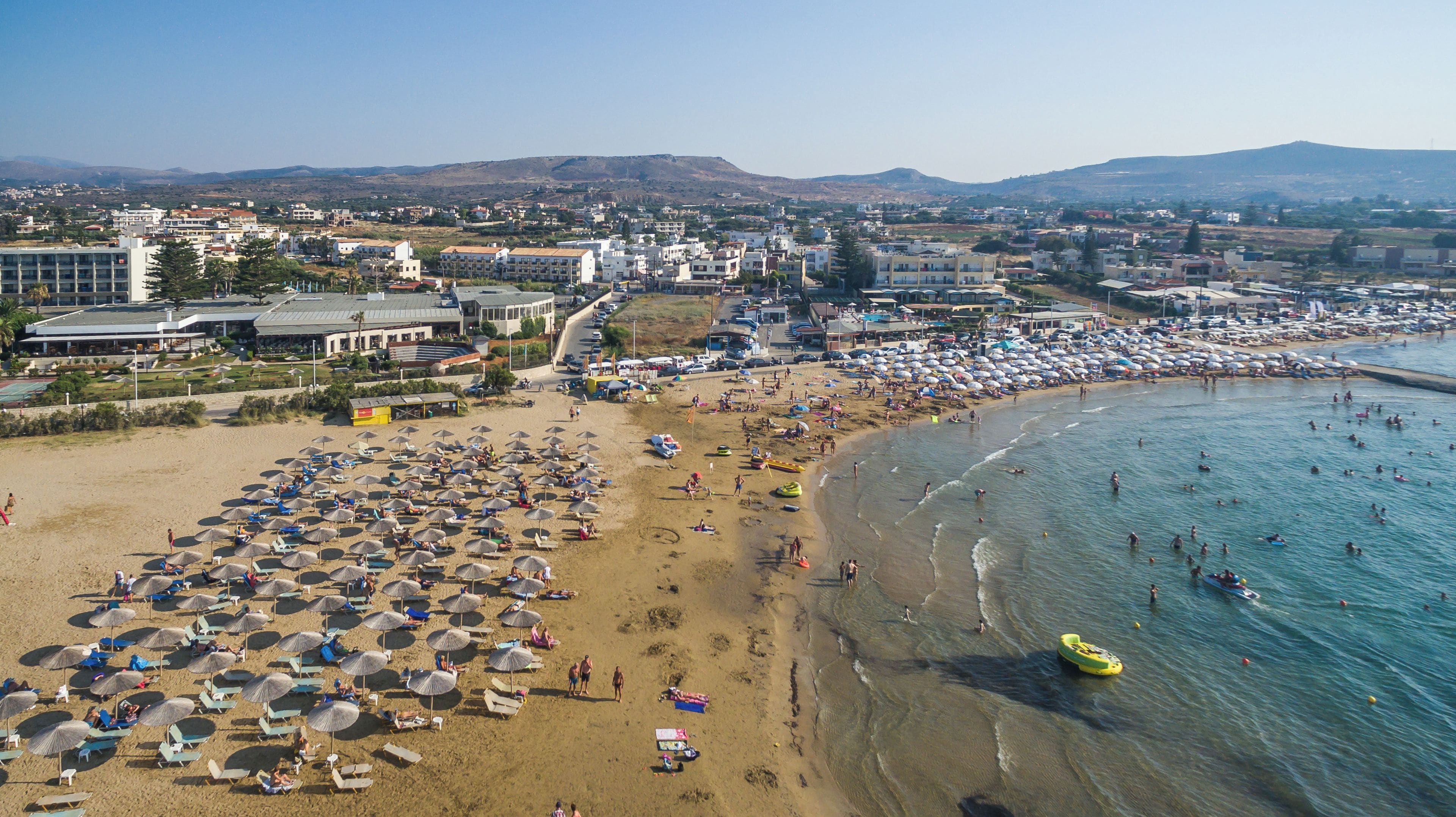 A beach with numerous umbrellas and sun loungers, a clear sky, and a mountainous backdrop.