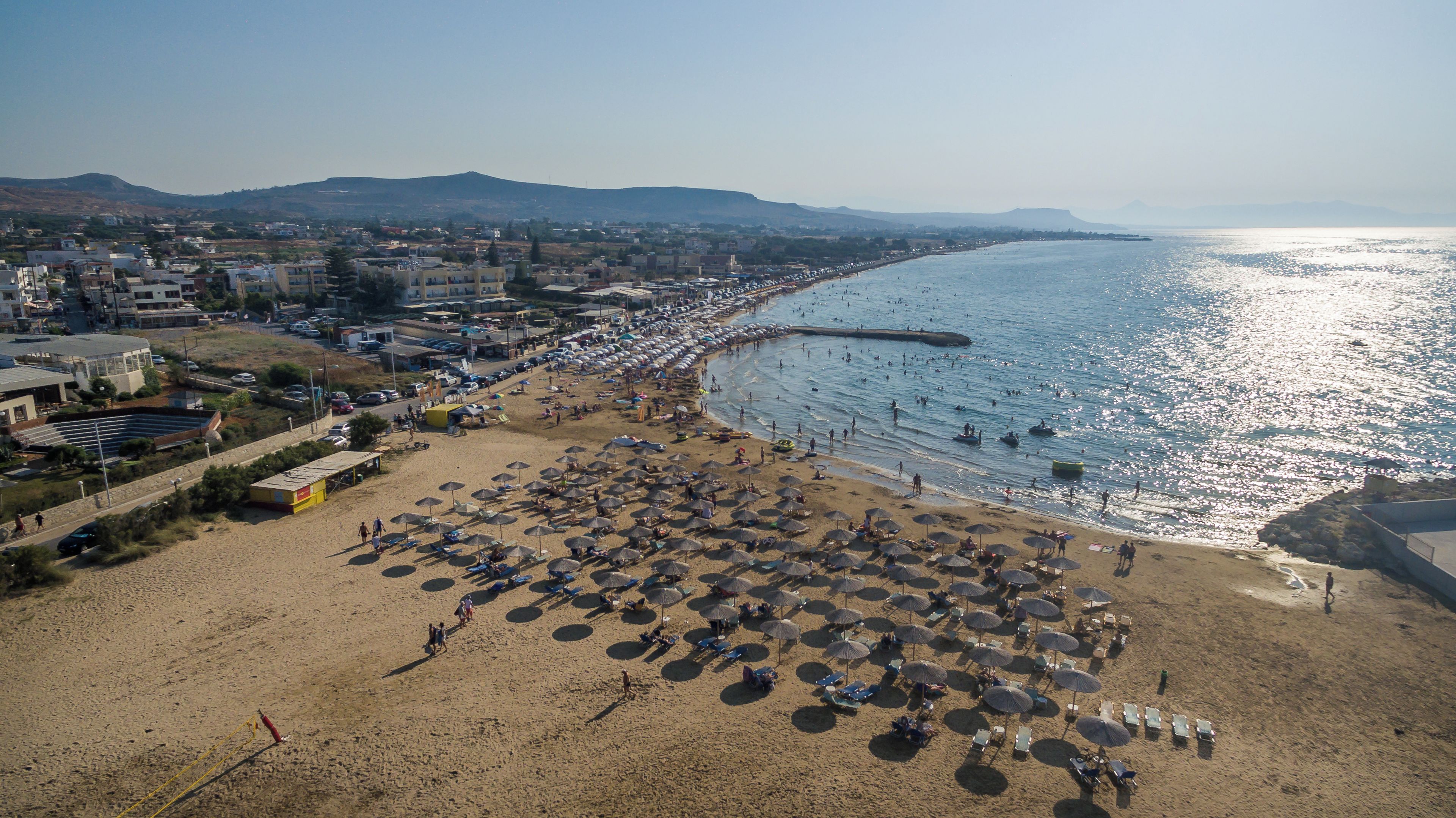 Ein Strand mit vielen Sonnenschirmen und Liegestühlen, einem klaren Himmel und einer Küstenlinie mit Gebäuden im Hintergrund.