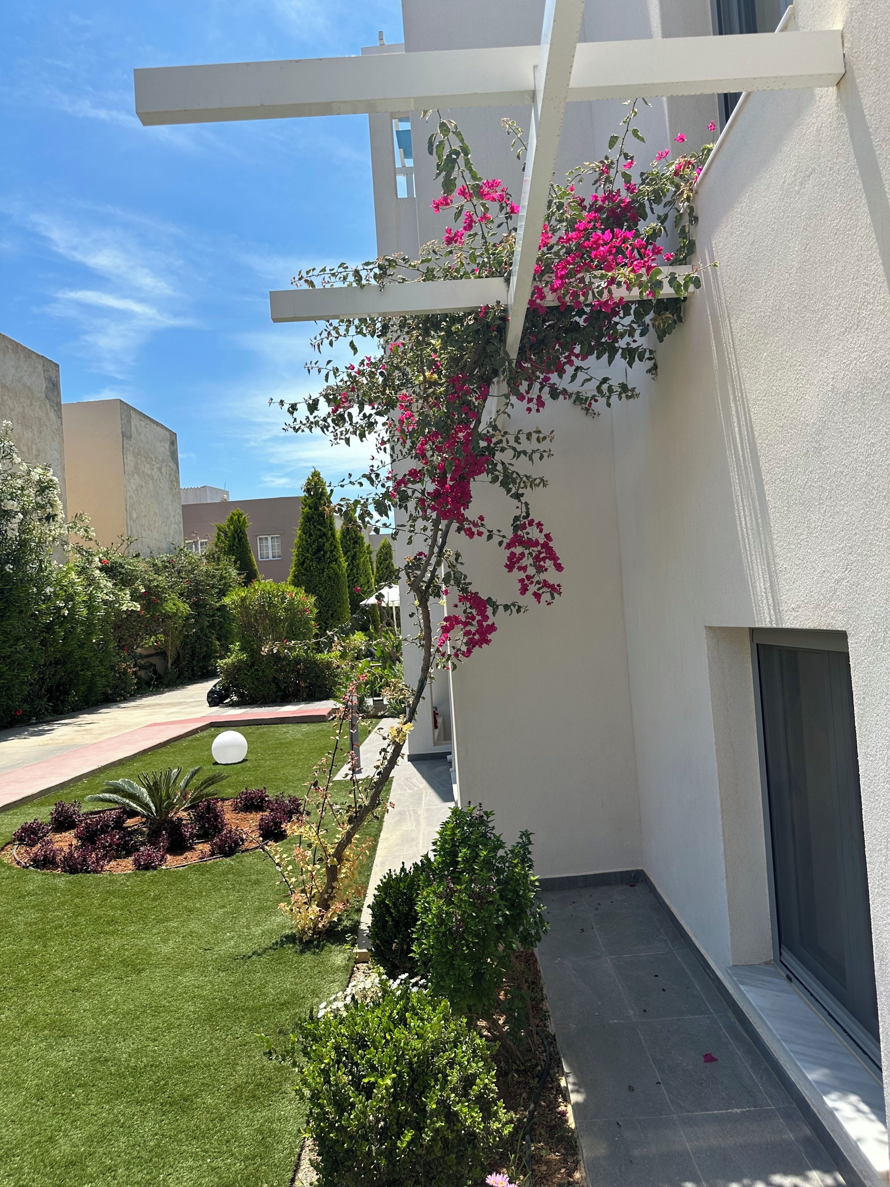 Un edificio moderno con facciata bianca, un balcone adornato da piante fiorite di colore rosa e un giardino curato con arbusti e prato.