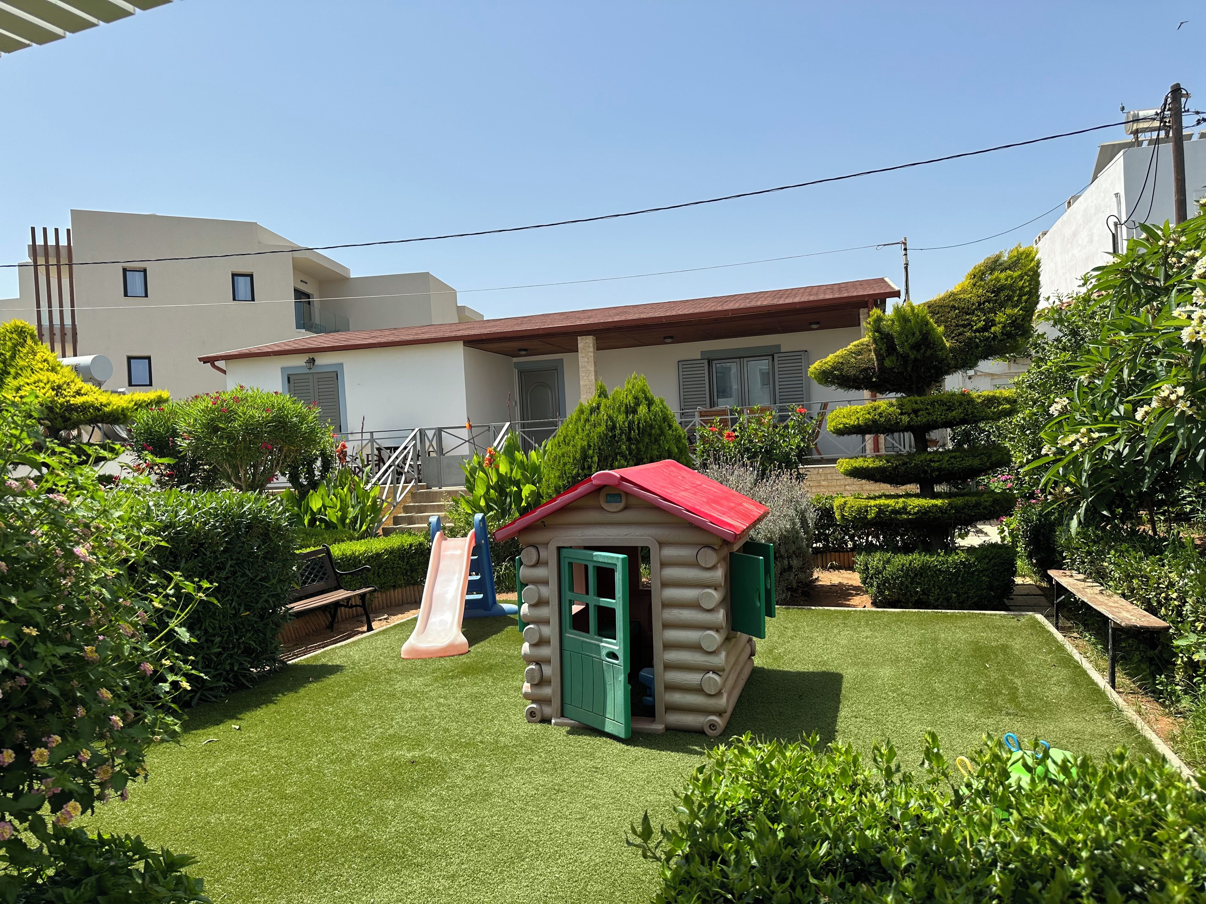 A small playhouse with a red roof and a green door, surrounded by a well-maintained lawn and various shrubs.
