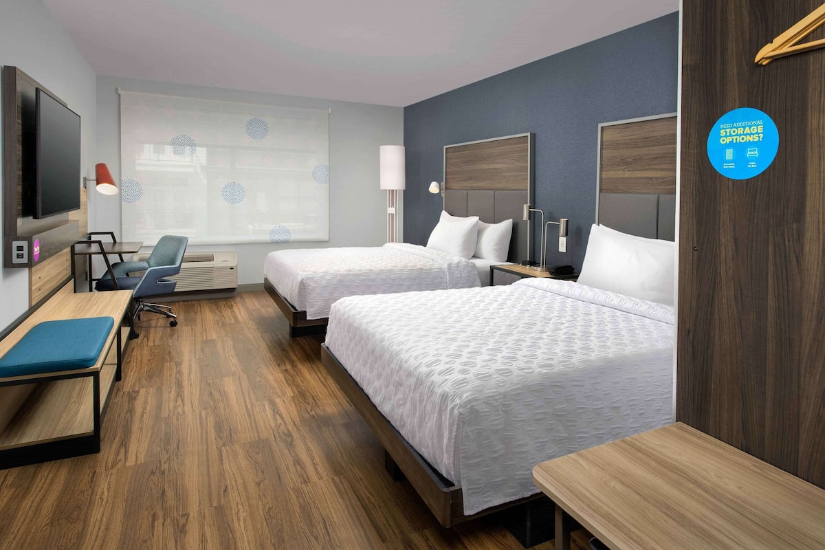 A hotel room with two beds, a desk, a chair, a TV, and a window with blinds.