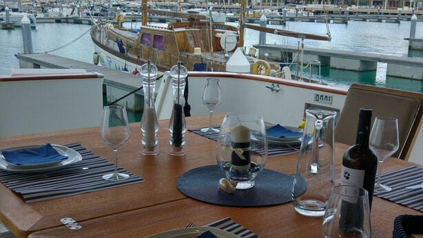 Ein Esstisch ist gedeckt mit Tellern, Gläsern und einer Flasche, mit Blick auf einen Hafen voller Boote und Gebäude.