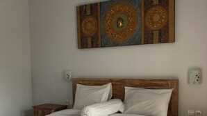 A neatly made bed with white linens and two folded towels, a wooden headboard, and a large decorative wall piece above it. - Cantika Guest House (Pecatu)