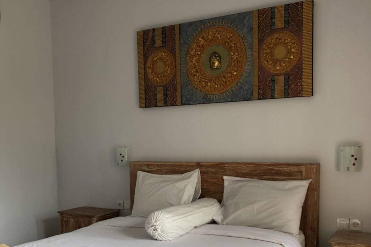 A neatly made bed with white linens and two folded towels, a wooden headboard, and a large decorative wall piece above it.