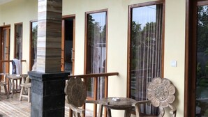 A covered outdoor seating area with wooden chairs and tables, a central pillar, and large windows with curtains. - Cantika Guest House (Pecatu)