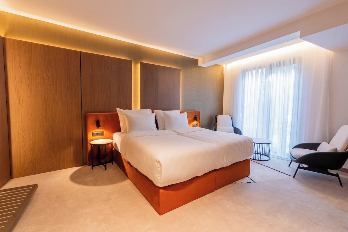 A modern hotel room with a large bed, a chair, a small table, and a window with curtains.