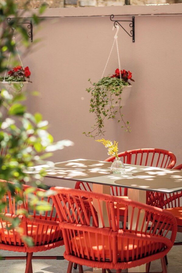 Una mesa redonda con un florero de vidrio, rodeada de sillas rojas, y macetas colgantes con flores rojas.