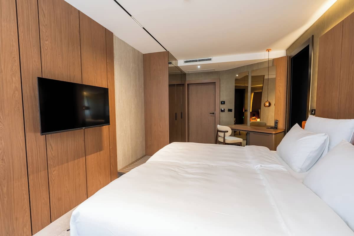 A modern hotel room with a large bed, a flat-screen TV mounted on the wall, a wooden wardrobe, a desk with a chair, and a mirror.