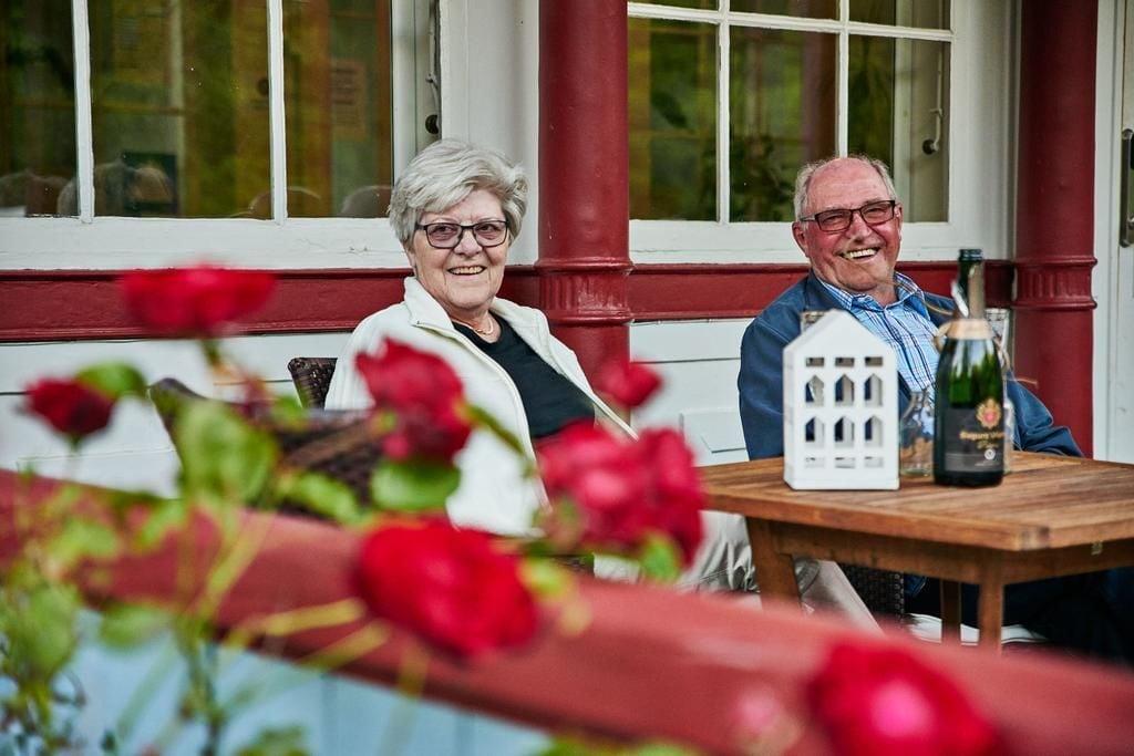 Two people seated at a wooden table with a bottle and a house-shaped object on it.