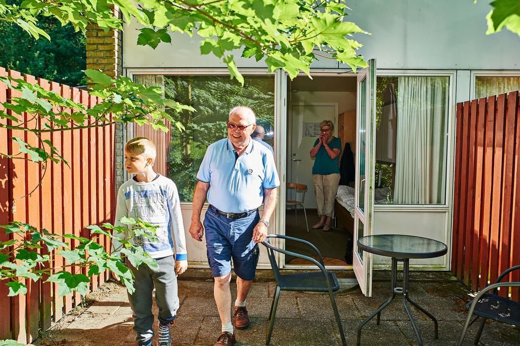 A man and a child walking towards a glass door, with a metal chair and a small table visible in the foreground.