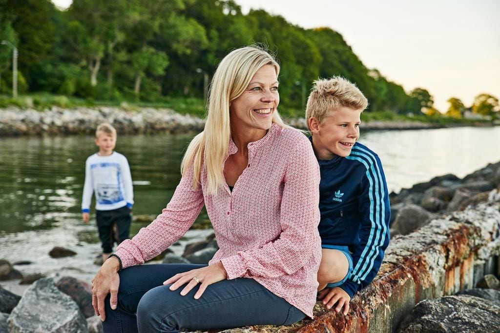 En person sidder på en vejrbidt stenvæg ved et vand, med grøn vegetation i baggrunden.