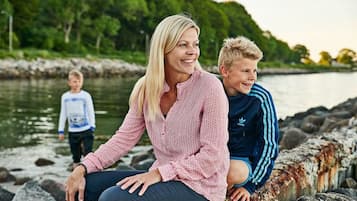 En person sidder på en vejrbidt stenvæg ved et vand, med grøn vegetation i baggrunden.