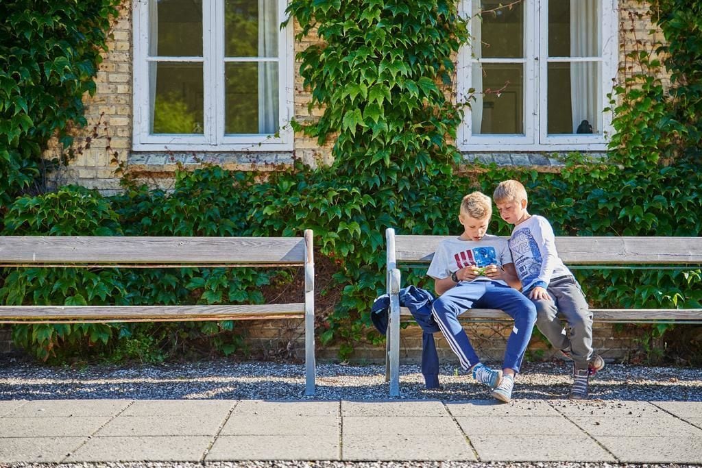 Deux enfants sont assis sur un banc à l’extérieur, devant un bâtiment en briques et sur fond de verdure.