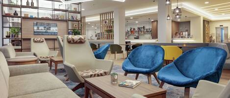 Un área de estar moderna con sofás, sillones y una mesa de centro. Hay estanterías con libros y objetos decorativos, y una cocina al fondo.