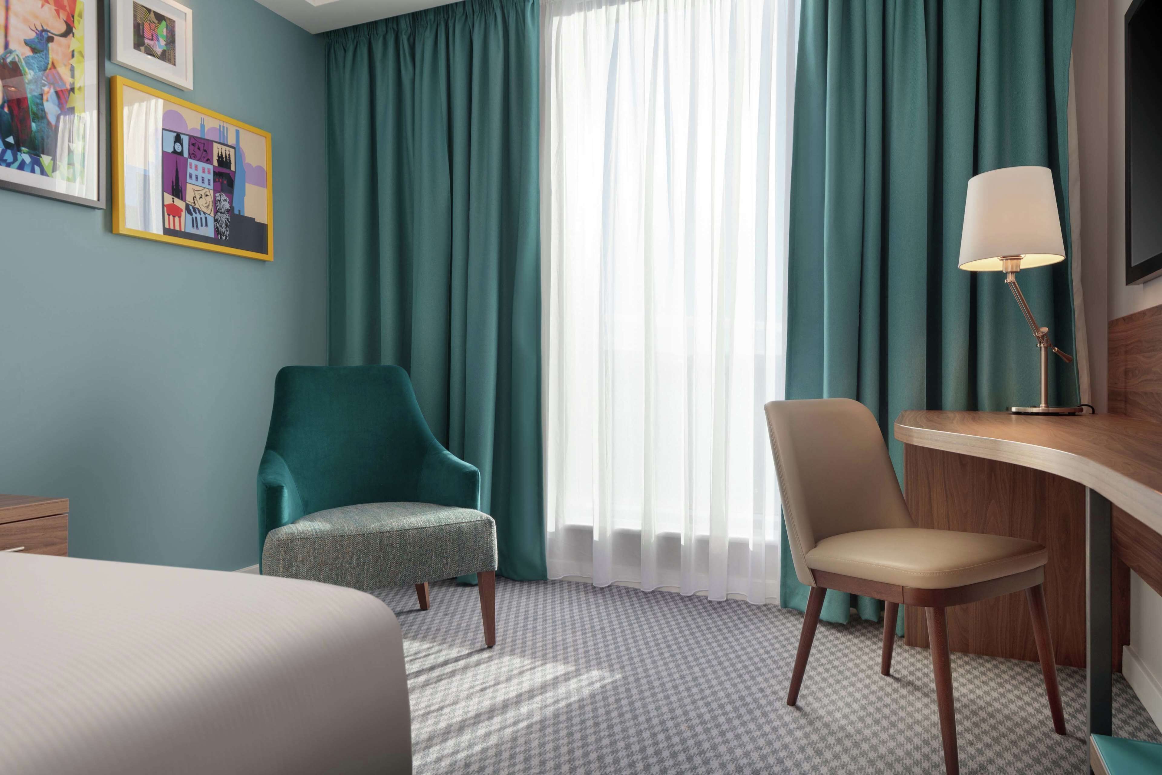 Une chambre d’hôtel avec des rideaux de couleur turquoise, un lit, un bureau avec une lampe et deux chaises.