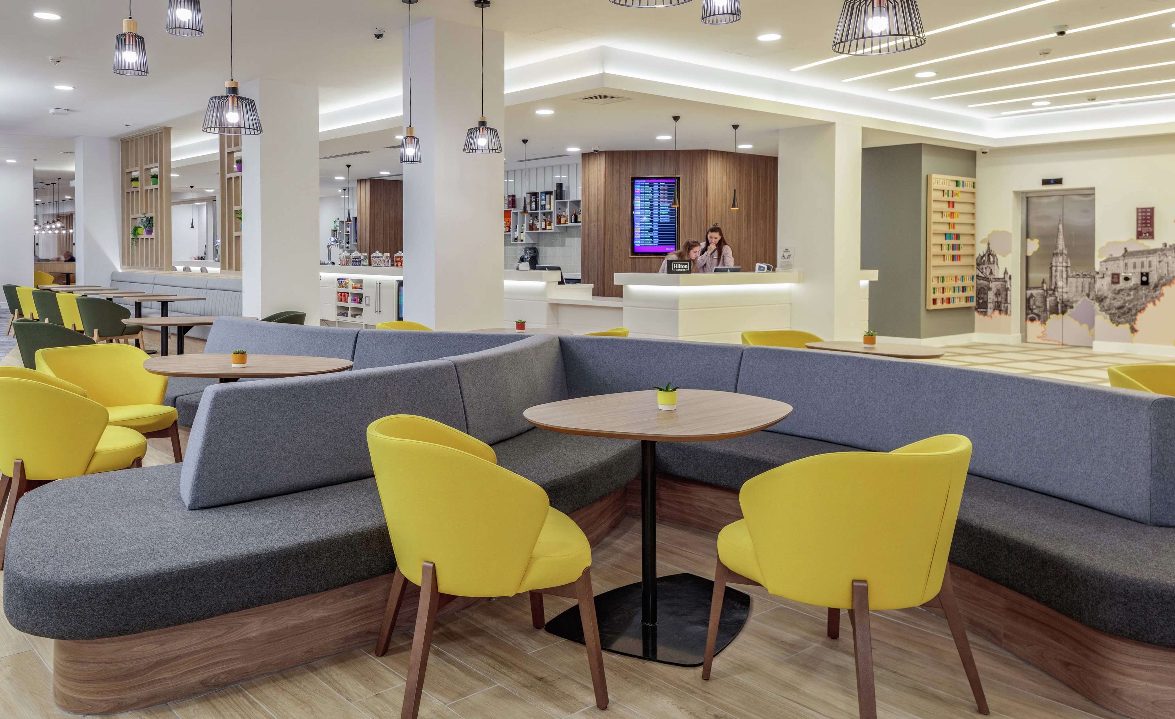 Un hall d’hôtel moderne avec un espace salon comprenant un canapé d’angle gris et des fauteuils jaunes, un comptoir de réception et un écran numérique.