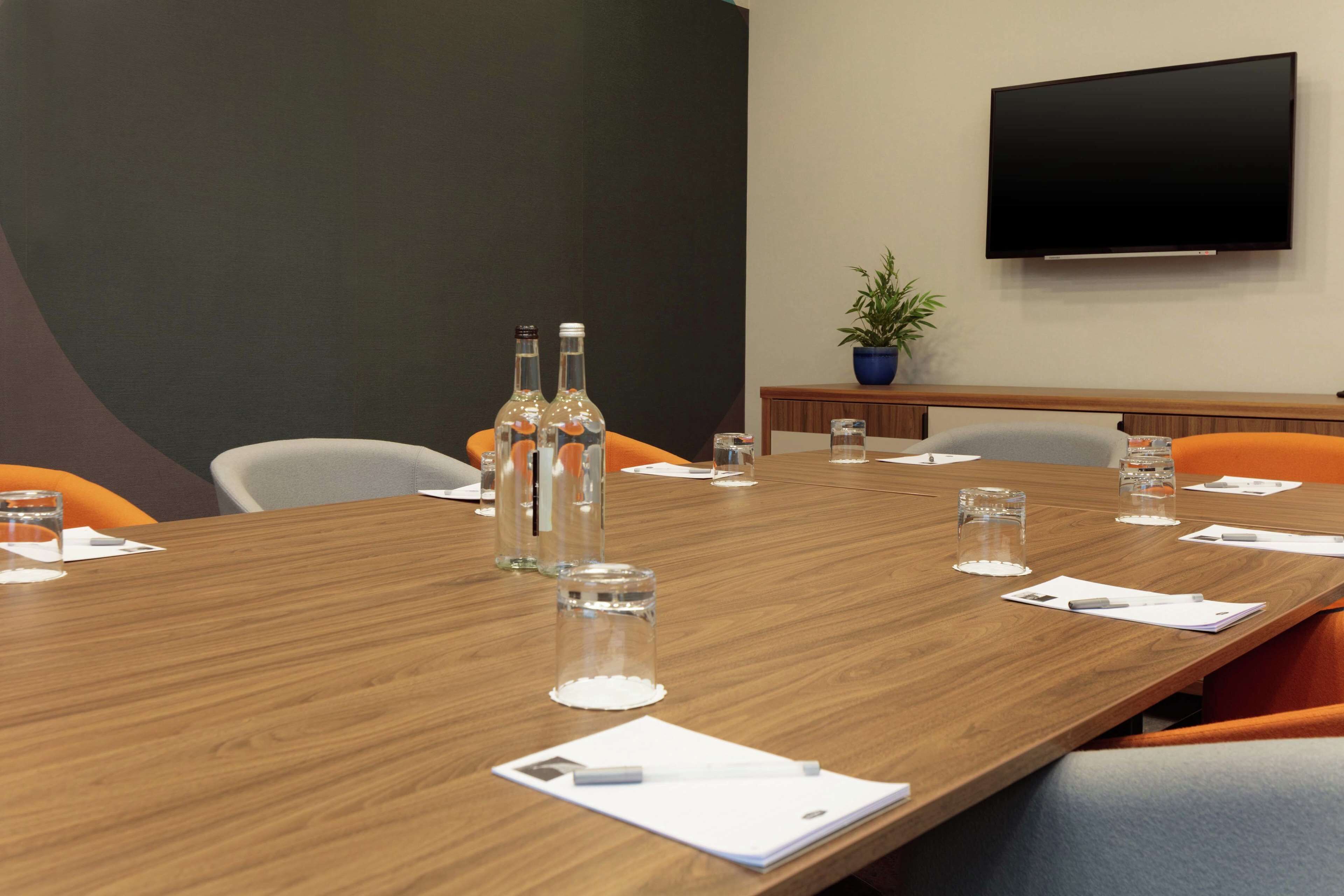 A conference room with a long wooden table, chairs, water bottles, glasses, and a flat-screen TV on the wall.