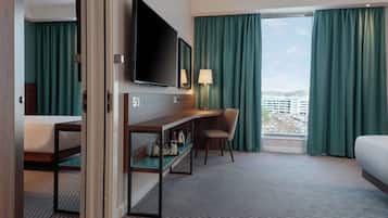 Et hotelværelse med seng, skrivebord med stol, fjernsyn og et vindue med gardiner.