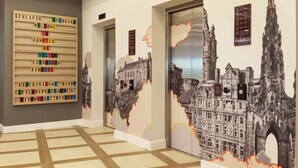 Un couloir d’hôtel avec une pancarte d’accueil, des ascenseurs et une grande fresque représentant un bâtiment historique.