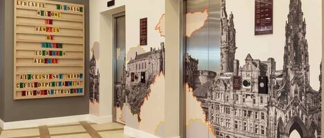 A hotel hallway with a welcome sign, elevators, and a large mural of a historic building.