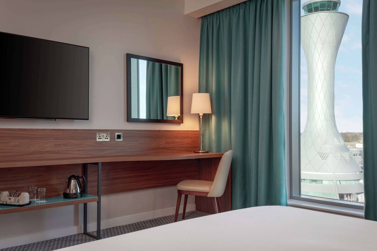 A hotel room with a bed, a desk with a chair, a television, a lamp, and a view of a unique building through a window.