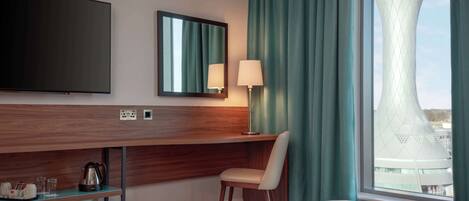A hotel room with a bed, a desk with a chair, a television, a lamp, and a view of a unique building through a window.