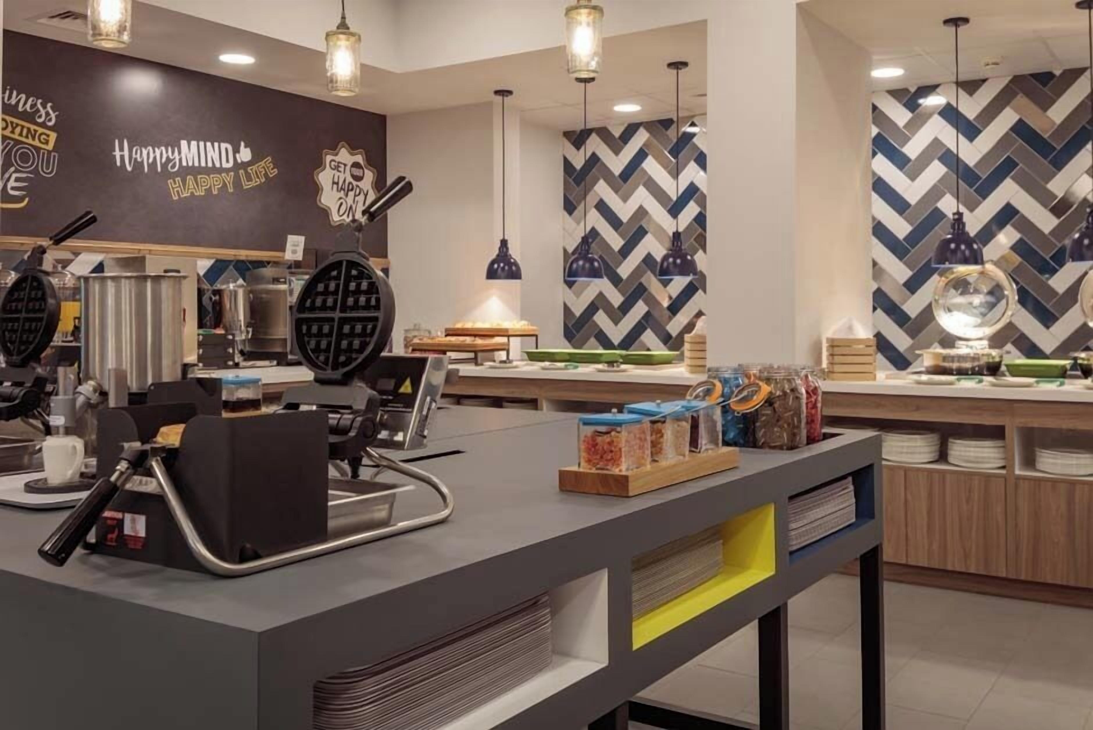 A modern cafe with a central counter, waffle maker, and various food items.