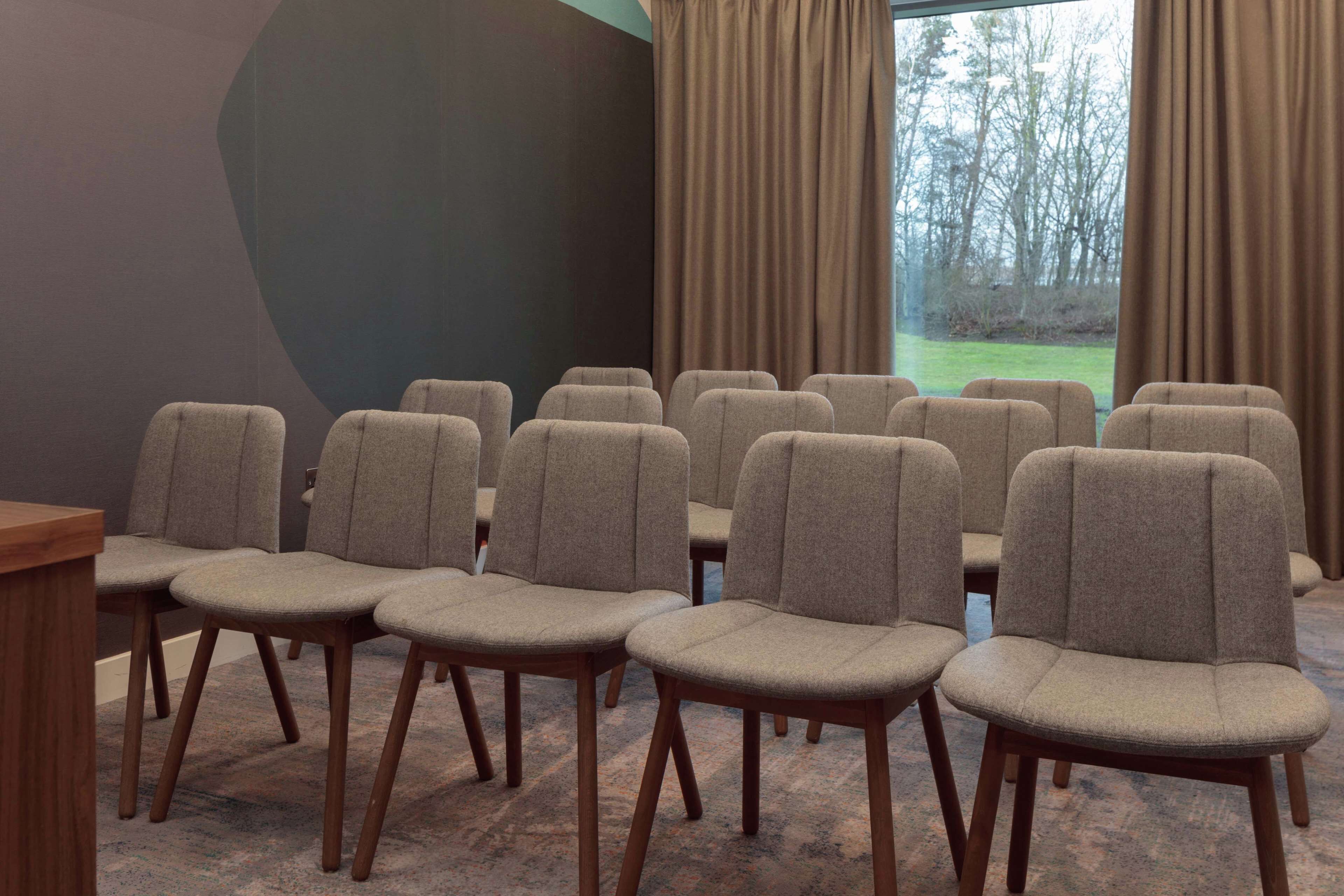 A conference room with rows of chairs and a table, large windows with curtains, and a view of trees outside.