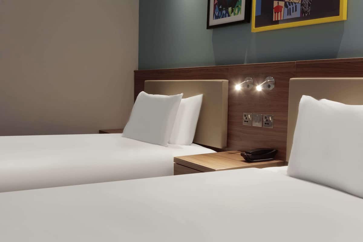 A hotel room with two beds, a wooden headboard, a bedside table with a phone, and wall-mounted light fixtures.