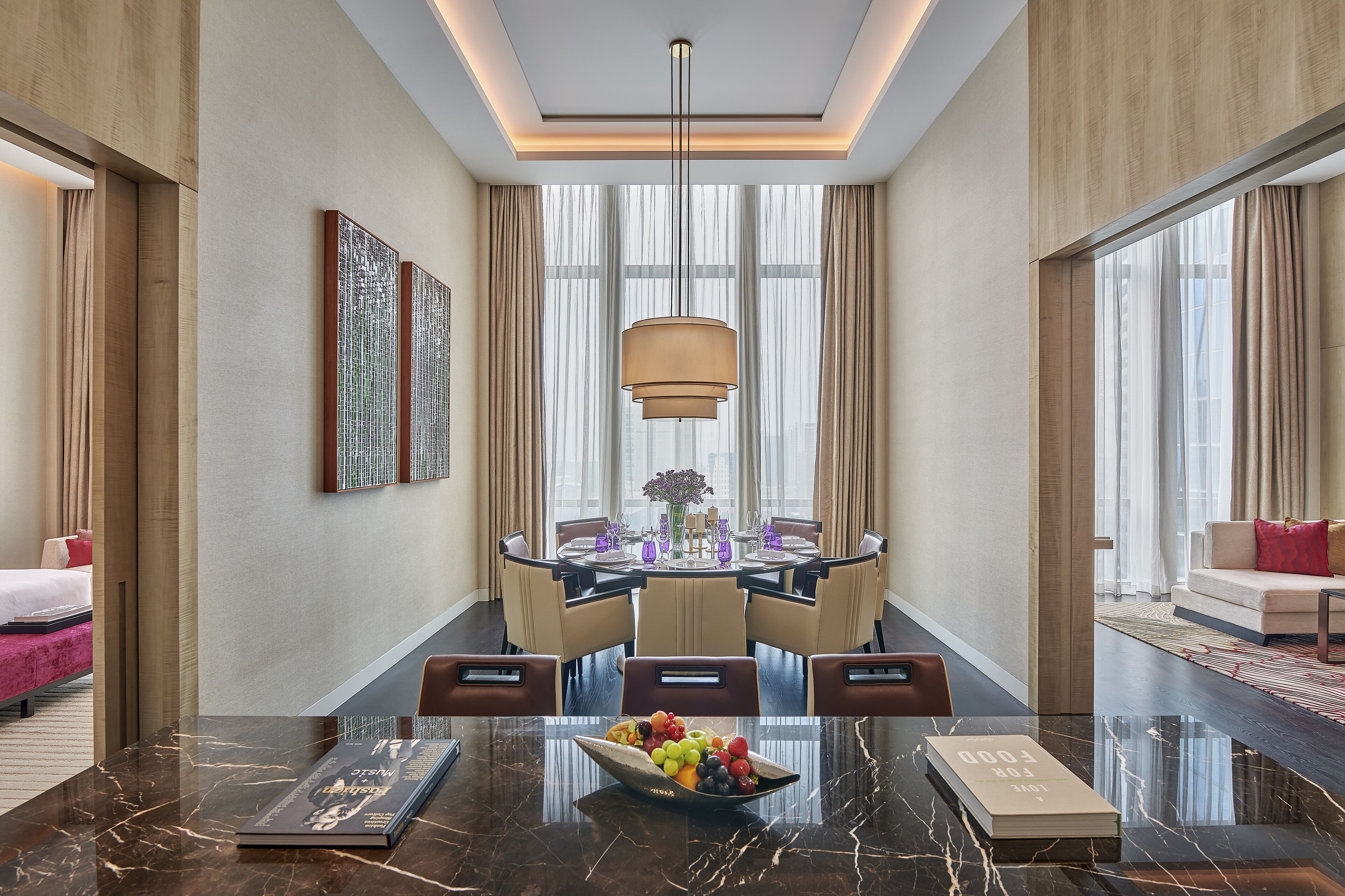 A modern hotel dining area with a marble table, a chandelier, and a view of the city through large windows.