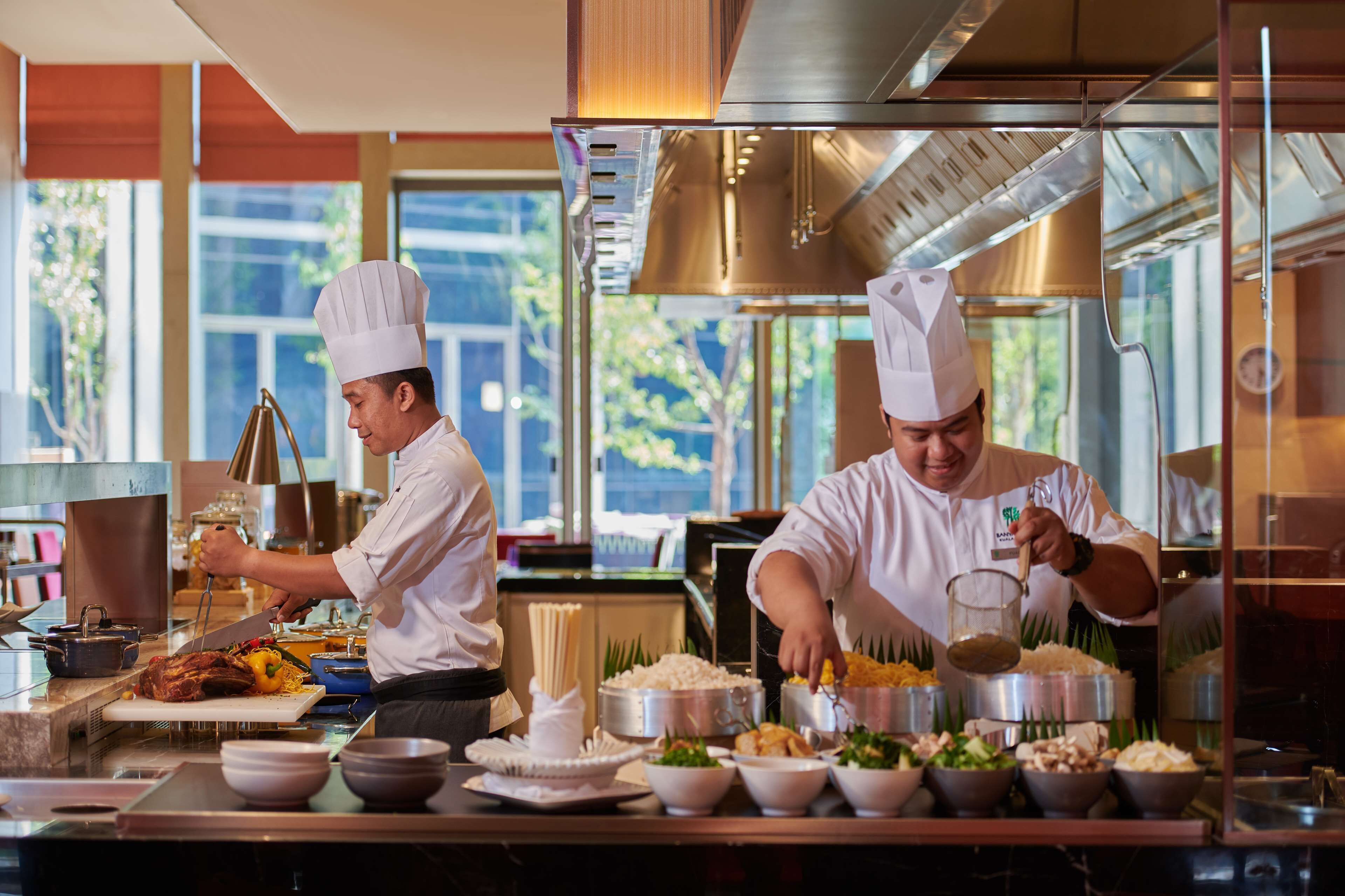 Two chefs preparing food in a modern kitchen with stainless steel appliances and large windows.