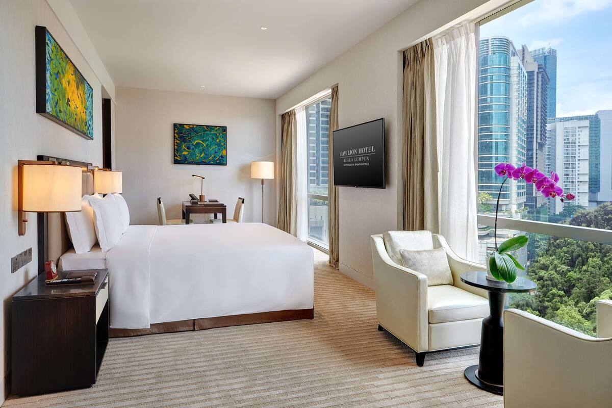 Une chambre d’hôtel avec un grand lit, deux tables de chevet avec des lampes, un canapé, une petite table avec une plante et une grande fenêtre donnant sur la ville.