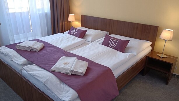 Una cama doble con sábanas blancas y detalles morados, dos mesitas de noche con lámparas, y una ventana con cortinas.