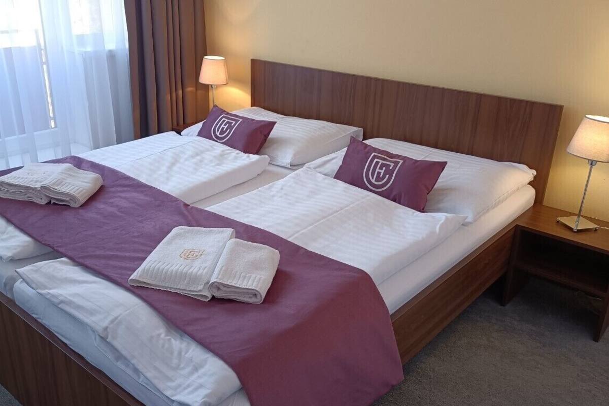 Una cama doble con sábanas blancas y detalles morados, dos mesitas de noche con lámparas, y una ventana con cortinas.