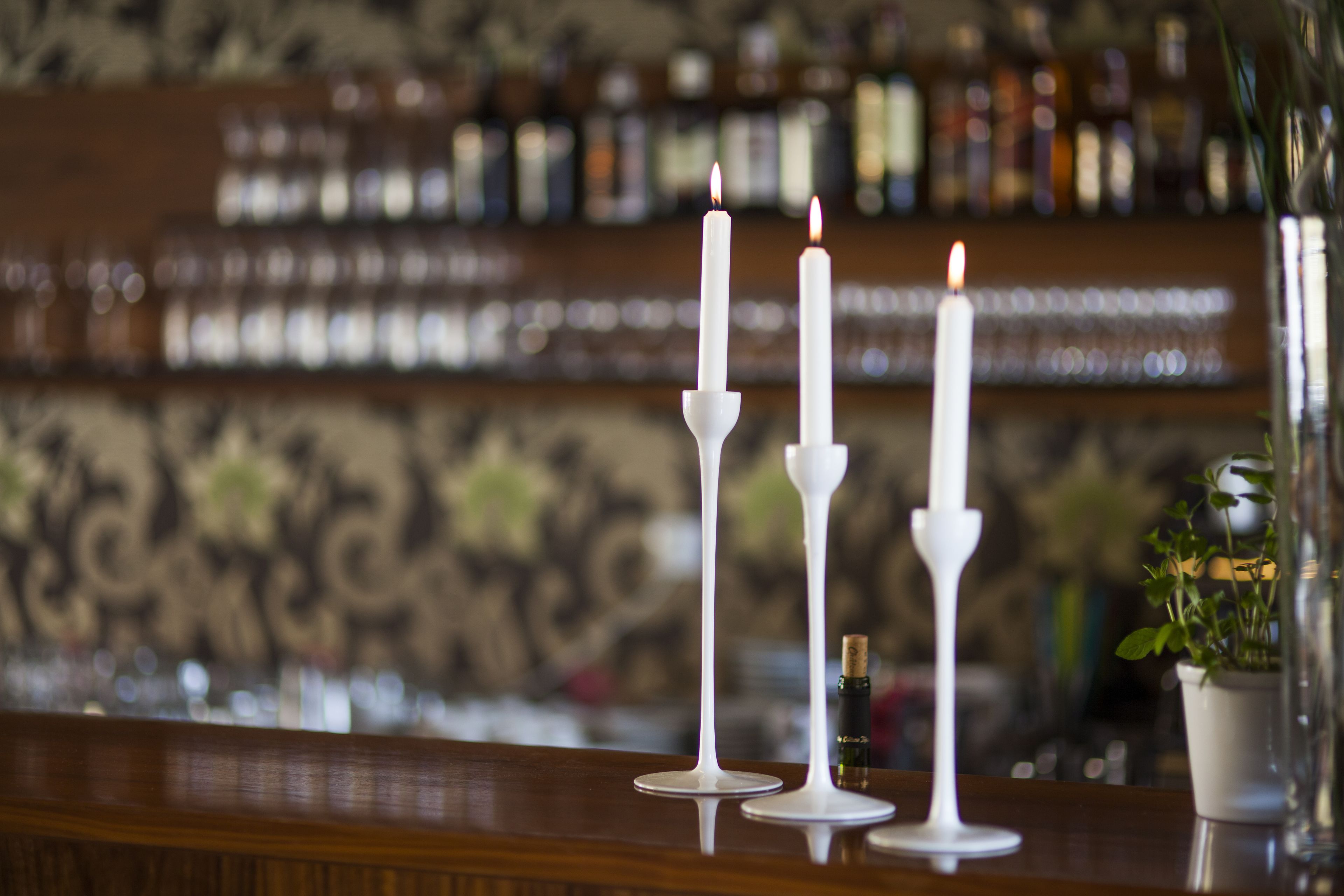Tres velas blancas sobre una barra, con un fondo borroso de estantes y botellas.