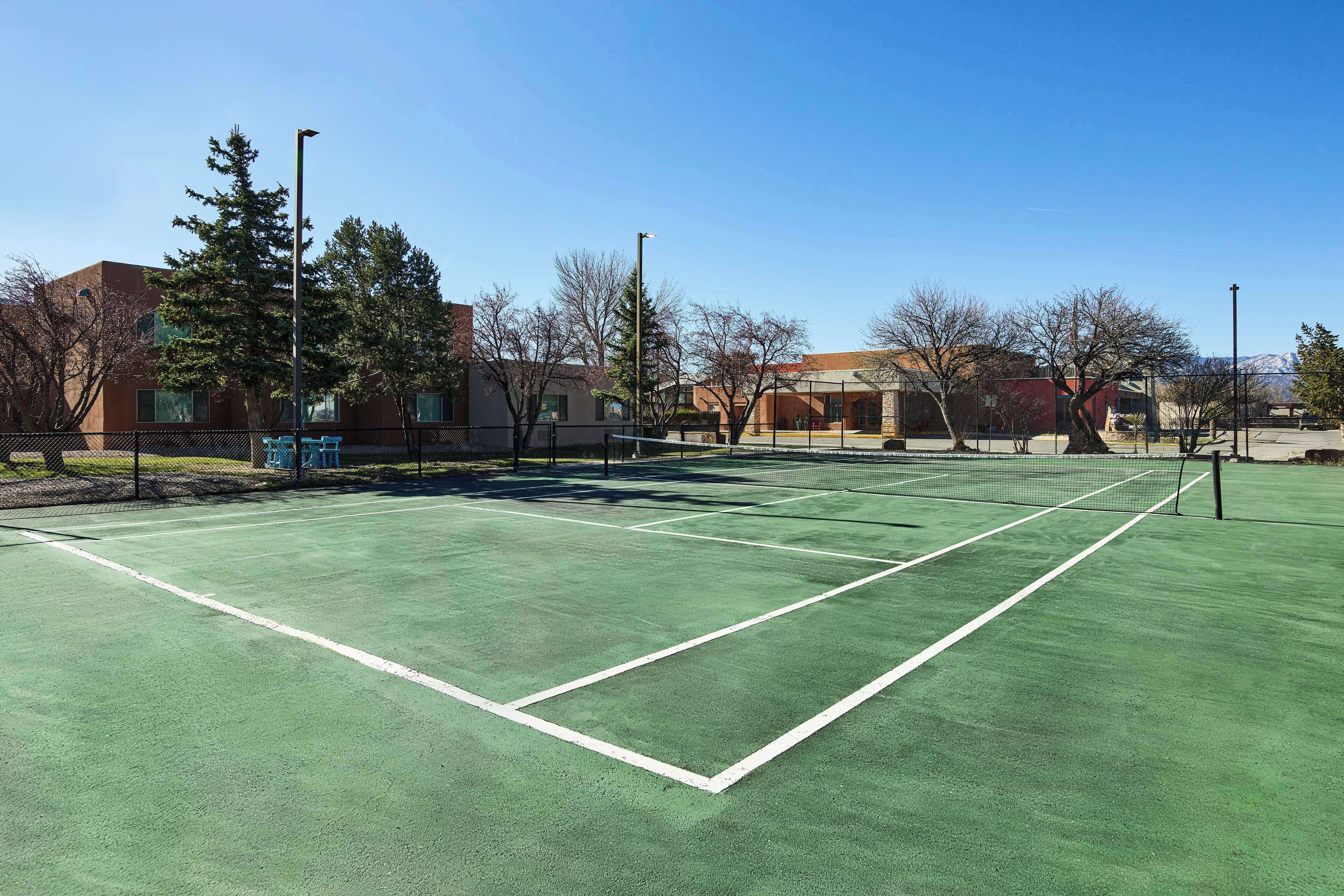 Un court de tennis avec une surface verte et des lignes blanches, entouré d’arbres et d’immeubles.