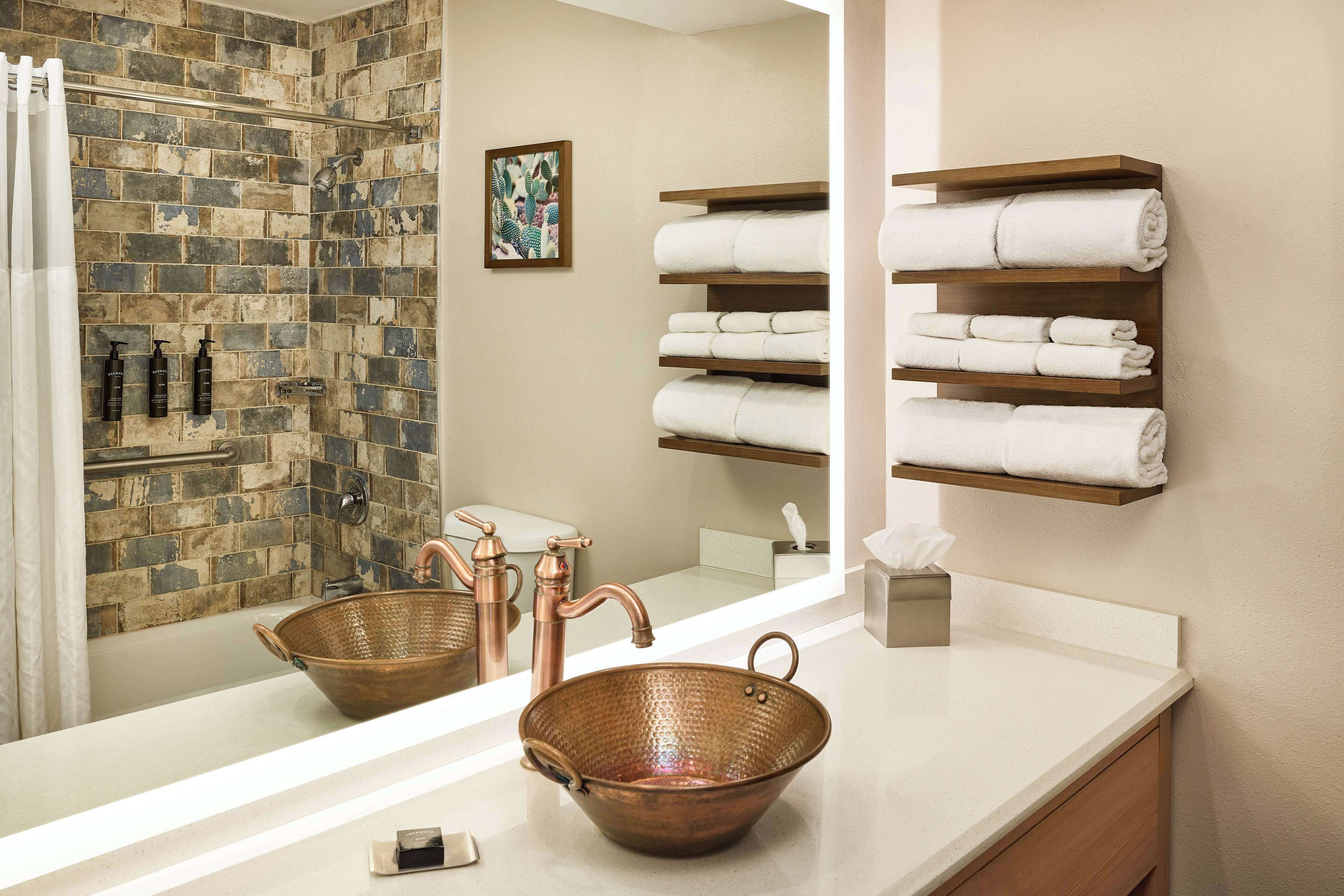 A bathroom with a copper basin, a large mirror, a shower with a brick wall, and shelves with folded towels.