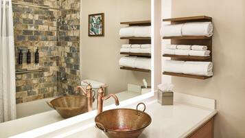 A bathroom with a copper basin, a large mirror, a shower with a brick wall, and shelves with folded towels.