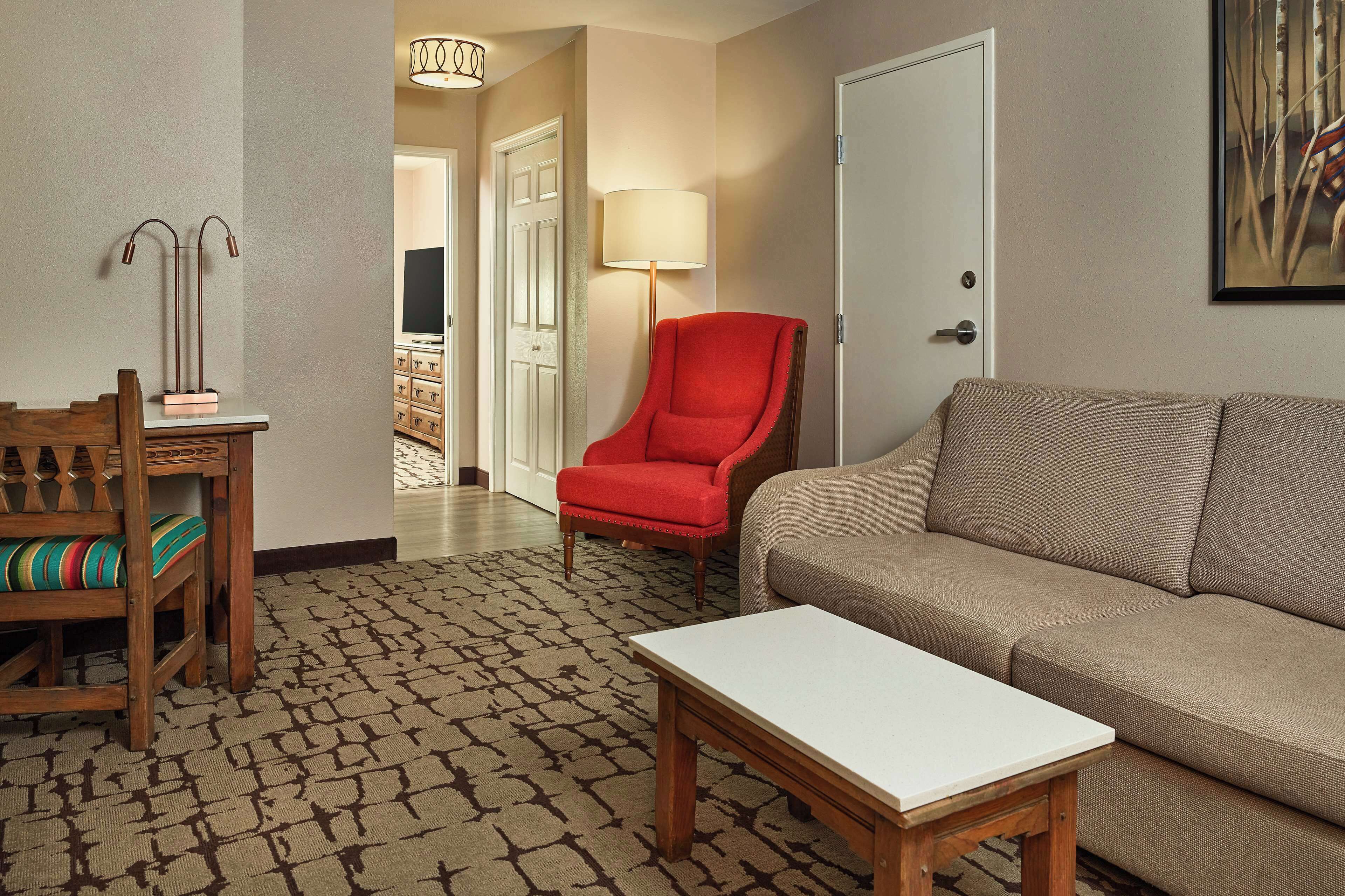 Une chambre d’hôtel avec une moquette à motifs, un bureau en bois avec une chaise, un fauteuil rouge, un canapé beige et une table blanche.