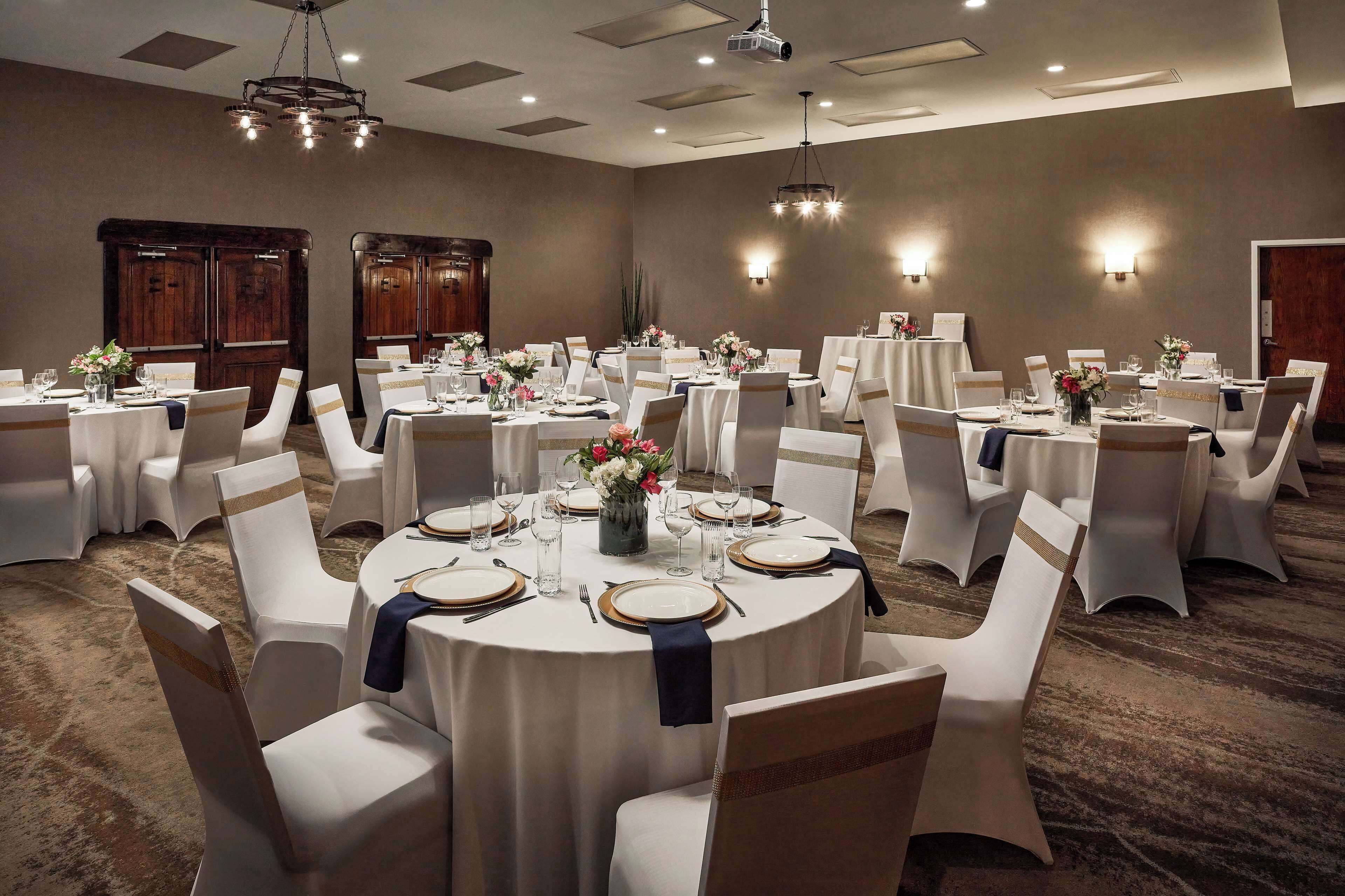 A banquet hall with round tables set for dining, each with white tablecloths, gold-rimmed plates, and white chairs with gold sashes.